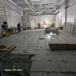 M&S GLOBAL SPÓŁKA Z OGRANICZONĄ ODPOWIEDZIALNOŚCIĄ - Układanie płytek w dużym pomieszczeniu z surowymi ścianami. Widoczne narzędzia i materiały budowlane. Dwie osoby pracują na rusztowaniu w tle.