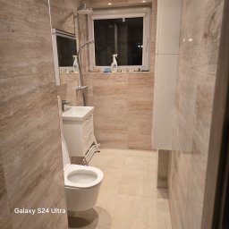 M&S GLOBAL SPÓŁKA Z OGRANICZONĄ ODPOWIEDZIALNOŚCIĄ - Beżowa łazienka po remoncie z toaletą, umywalką i kabiną prysznicową. Na parapecie okna stoją środki czystości. Widok z perspektywy wejścia.