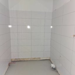 M&S GLOBAL SPÓŁKA Z OGRANICZONĄ ODPOWIEDZIALNOŚCIĄ - Wykończona łazienka z białymi, błyszczącymi płytkami na ścianach i szarą wylewką na podłodze. Widoczne rury i wystające kable elektryczne.