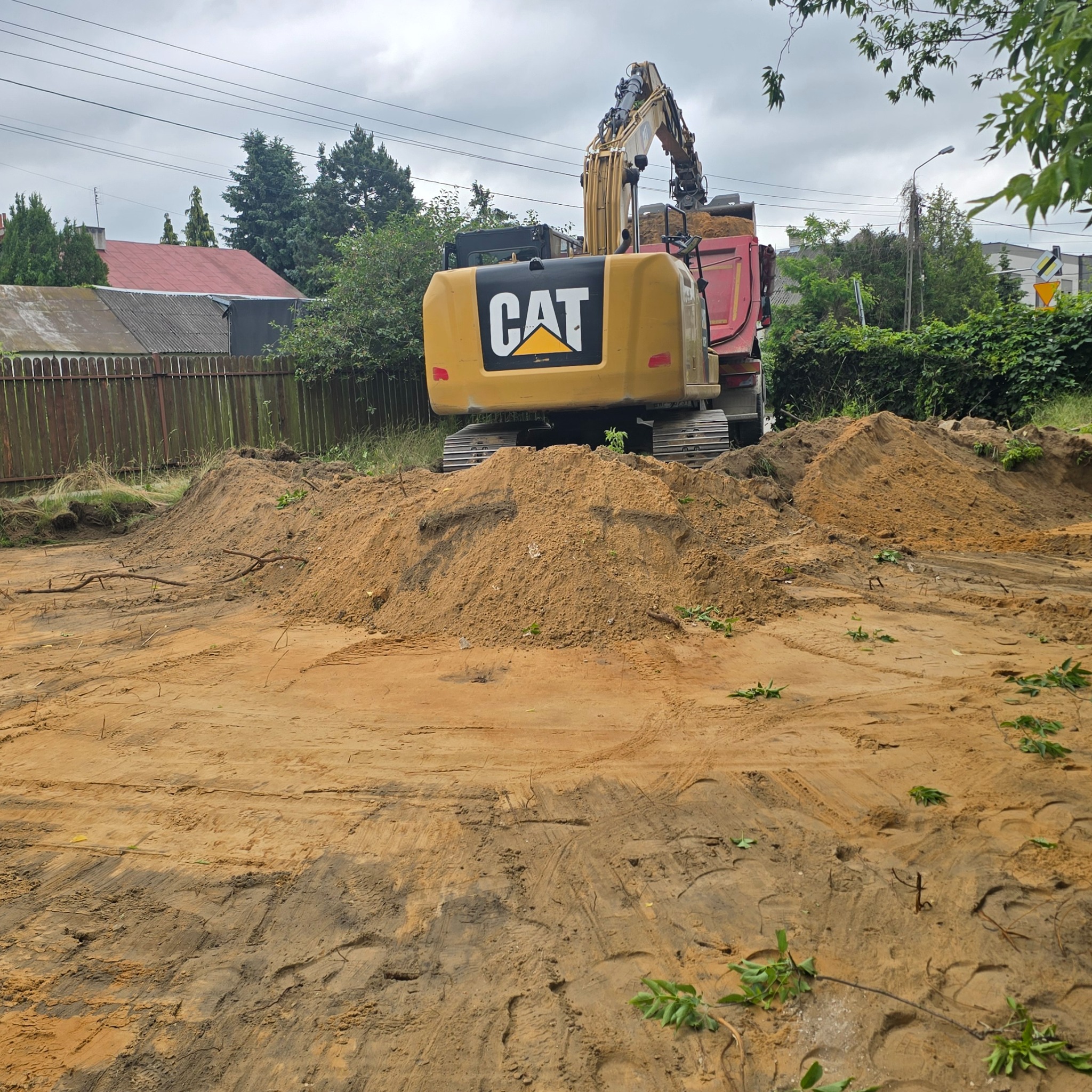 Żółta koparka CAT na placu budowy, widok z tyłu. Na ziemi świeża ziemia, w tle zieleń i budynki pod pochmurnym niebem.