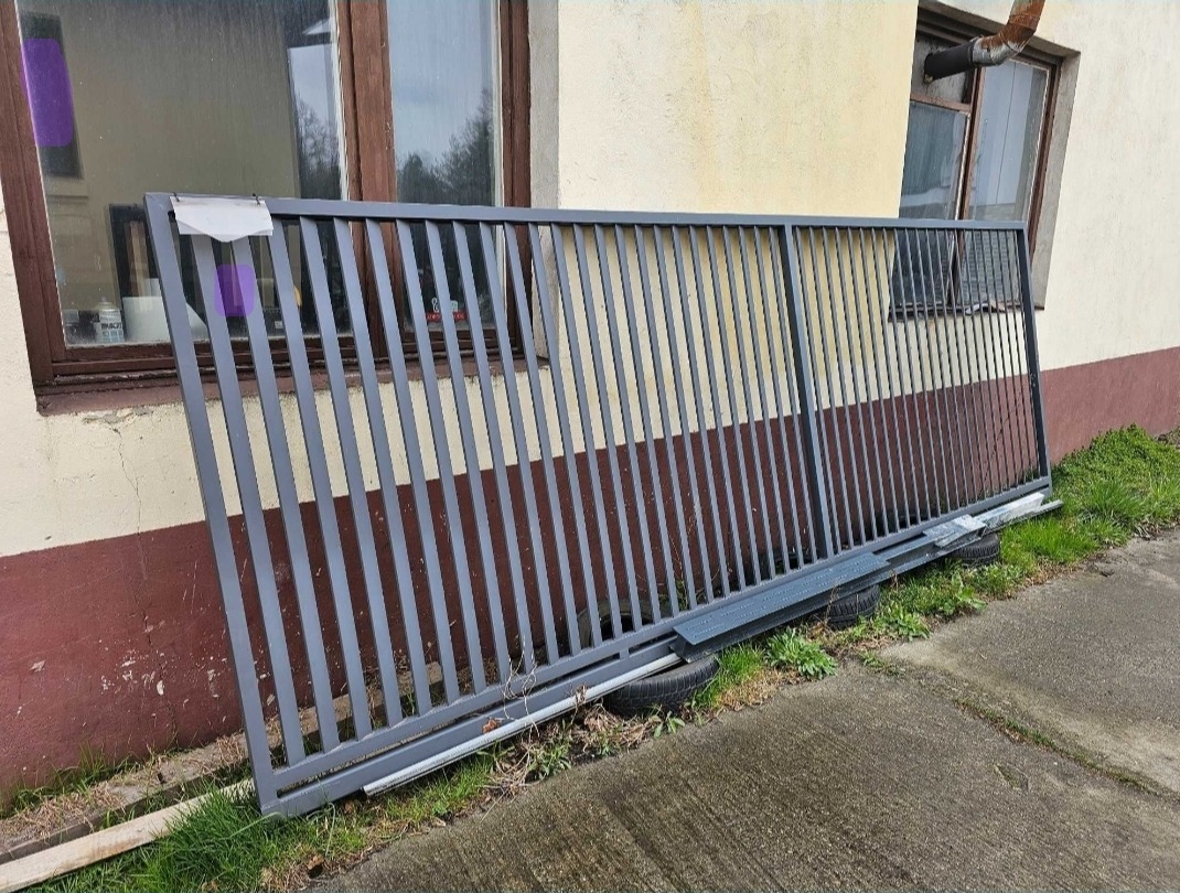 Szara, metalowa brama oparta o ścianę budynku z dwoma oknami. Brama leży na oponach. Widoczne fragmenty trawy i betonowej nawierzchni.