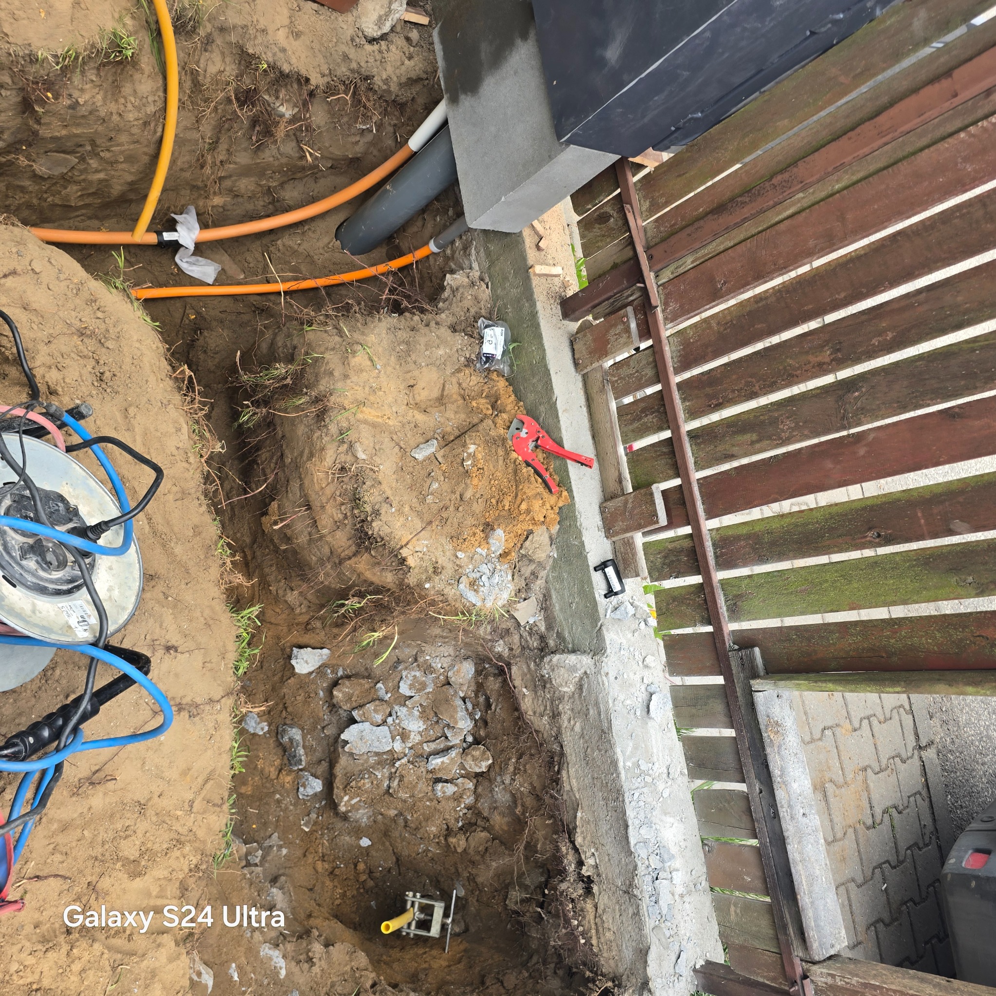 Wykop z rurami instalacyjnymi (pomarańczowe i szare) w ziemi, obok fundamentu i drewnianego płotu. Widoczne narzędzia i kable. Prace instalacyjne w toku.