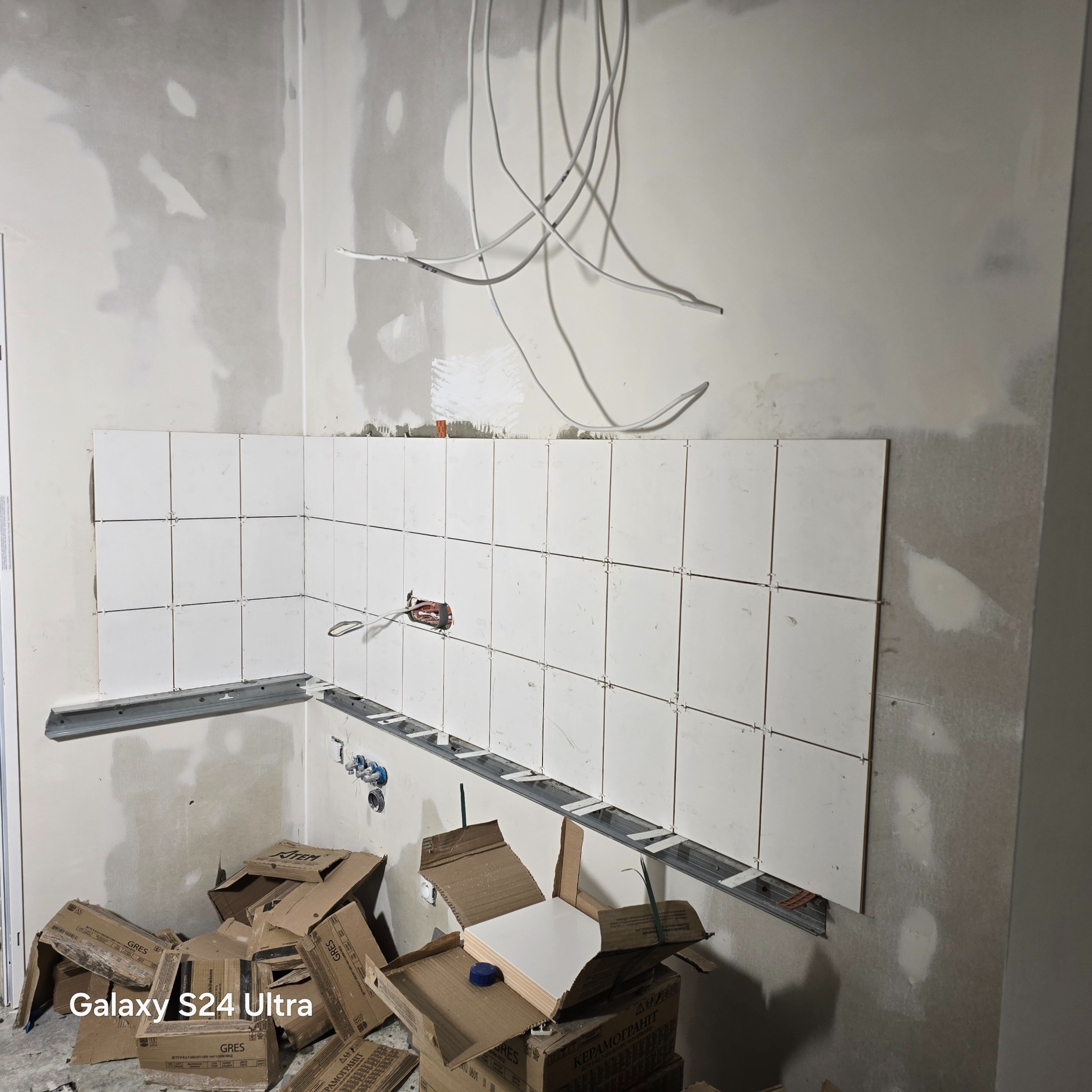 Układanie białych płytek w rogu pomieszczenia, widoczne metalowe profile i kartony po płytkach. Instalacja elektryczna na ścianie, etap prac wykończeniowych.