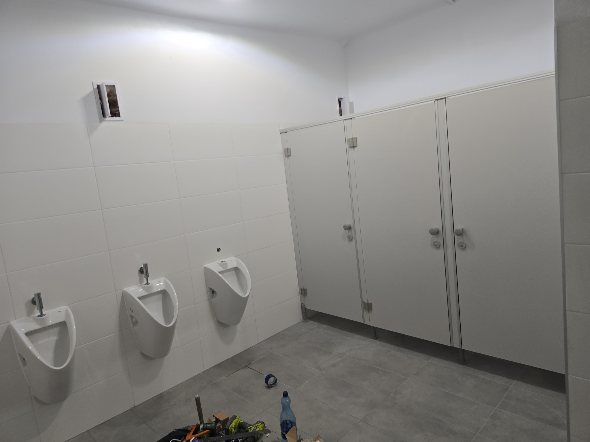 Publiczna toaleta w trakcie remontu: zamontowane pisuary i kabiny WC, widoczne narzędzia i materiały budowlane na szarej podłodze z płytek.