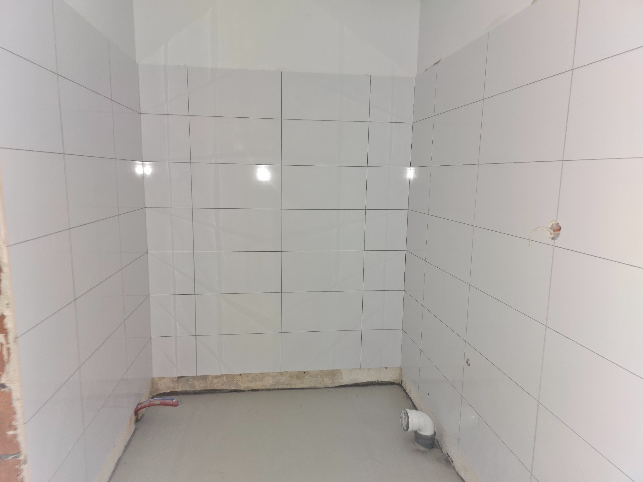 Wykończona łazienka z białymi, błyszczącymi płytkami na ścianach i szarą wylewką na podłodze. Widoczne rury i wystające kable elektryczne.