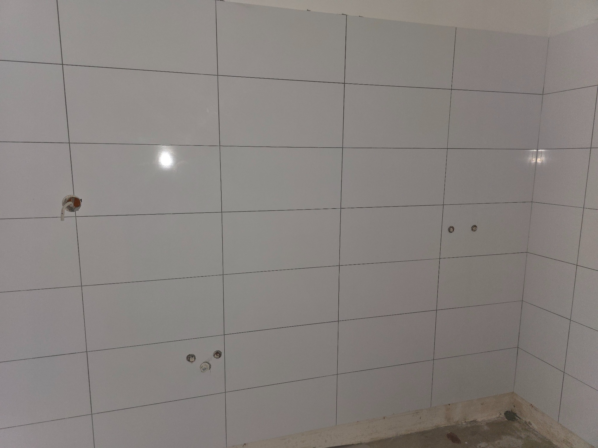 Wykafelkowana ściana w jasnych, kwadratowych płytkach z widocznymi otworami na instalacje. Świeżo położone kafelki na ścianie.