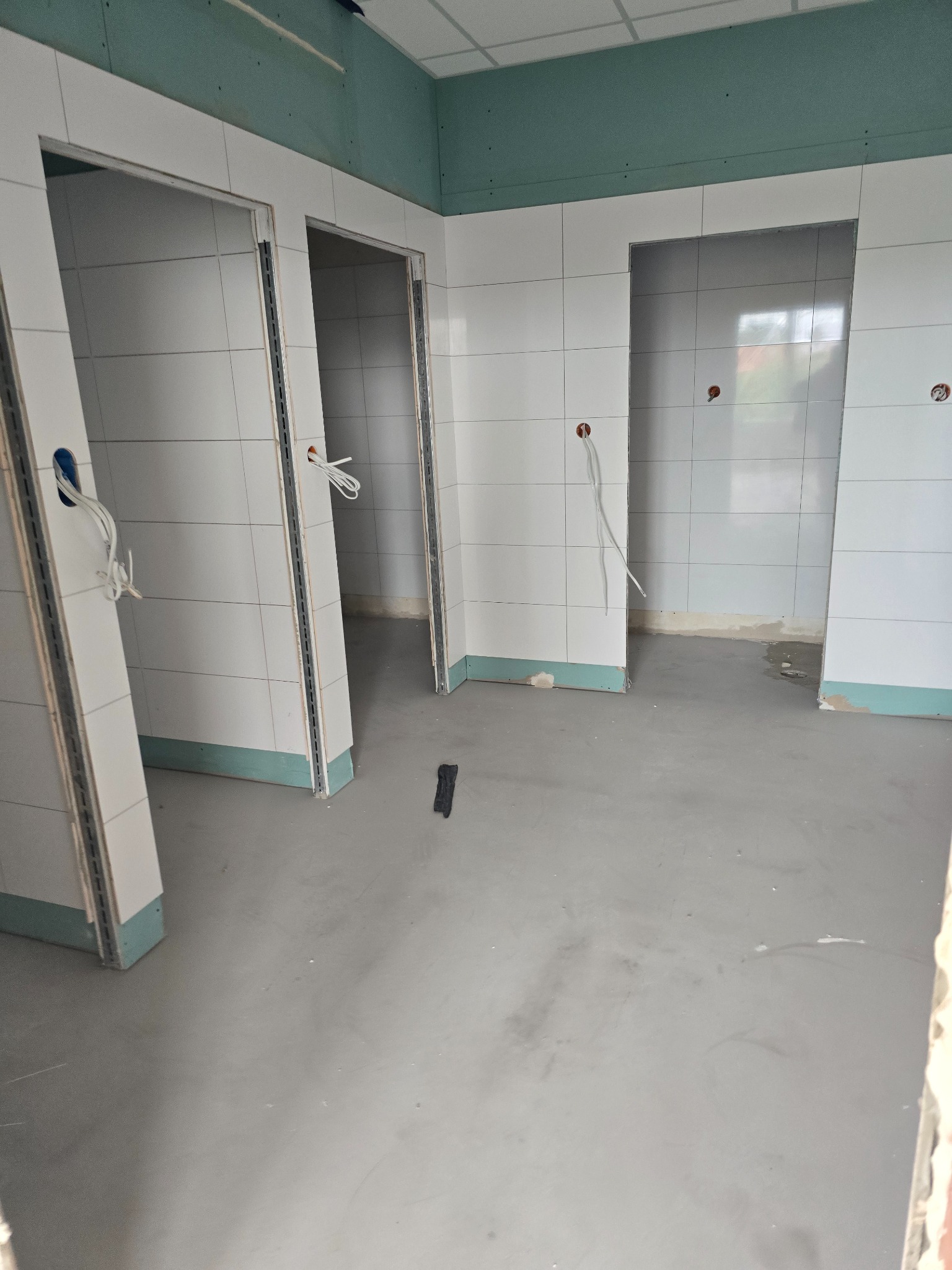 Wnętrze w trakcie remontu: białe płytki na ścianach, surowy beton na podłodze, widoczne otwory drzwiowe z metalowymi profilami. Instalacja elektryczna w przygotowaniu.