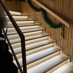 Drewniane schody z podświetleniem LED na każdym stopniu, udekorowane świątecznym wieńcem. Ciepłe światło podkreśla naturalną fakturę drewna.