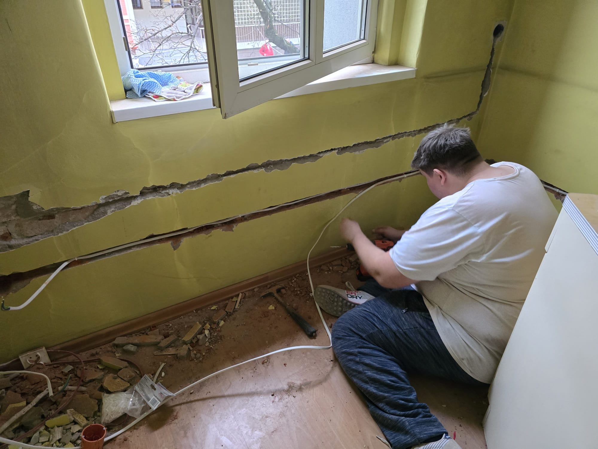 Elektryk w trakcie kucia bruzd pod instalację elektryczną w pokoju z żółtymi ścianami. Widoczne kable, narzędzia i gruz. Otwór w ścianie pod sufitem.