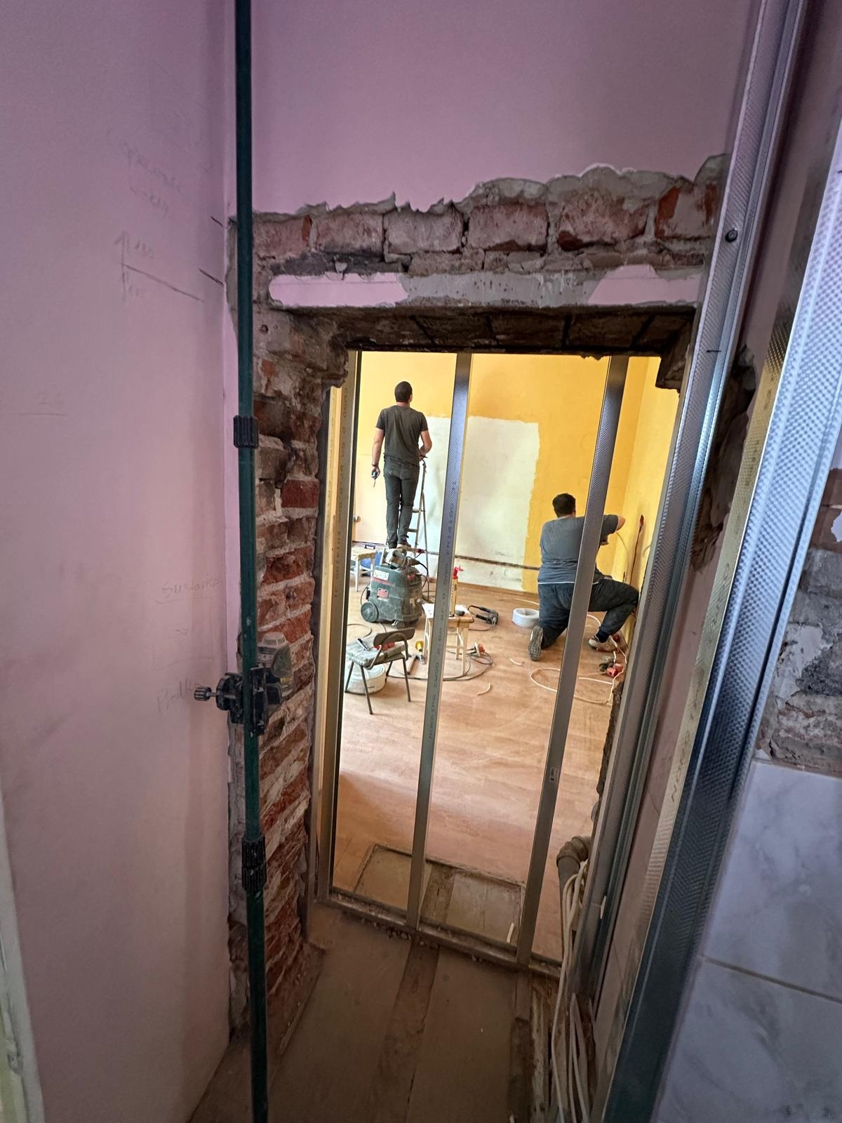 Remont wnętrza w toku: dwaj mężczyźni pracują w żółtym pokoju, jeden na drabinie, drugi przy ścianie. Widoczne narzędzia, instalacje i fragmenty konstrukcji. Ściany w trakcie wykończenia.