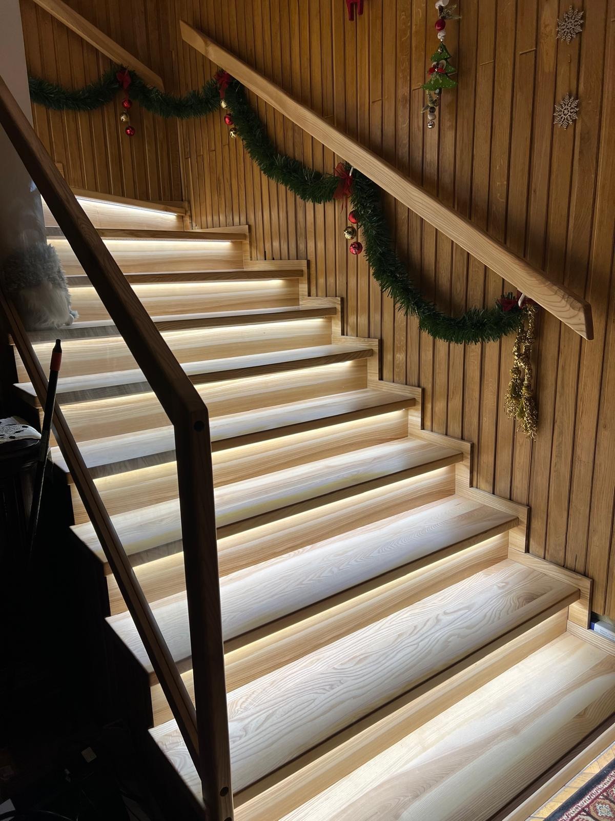 Drewniane schody z podświetleniem LED na każdym stopniu, udekorowane świątecznym wieńcem. Ciepłe światło podkreśla naturalną fakturę drewna.