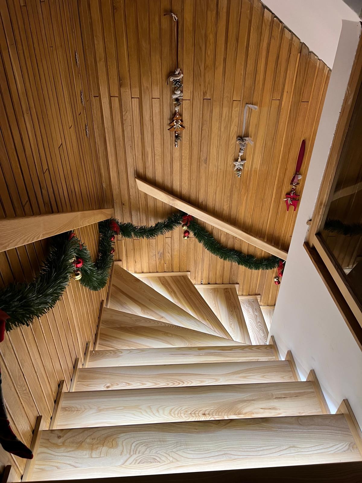 Drewniane schody z podświetleniem LED, ozdobione świąteczną girlandą. Widok z góry, ukazujący spiralny bieg schodów i drewnianą boazerię na ścianach i suficie.