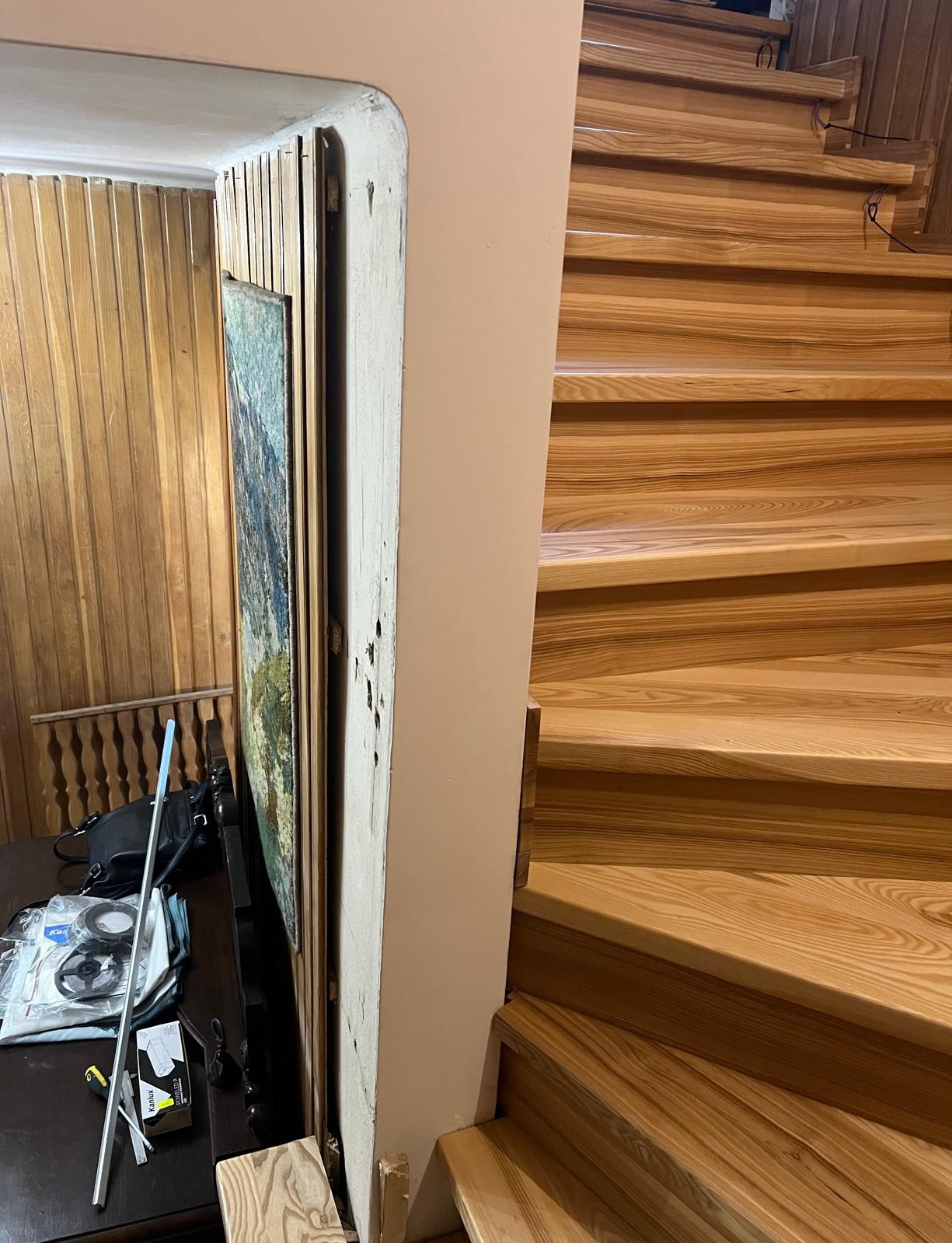 Drewniane schody z oświetleniem LED w trakcie montażu, widoczne przewody i elementy montażowe, częściowo widoczny obraz na ścianie, ciepłe oświetlenie.