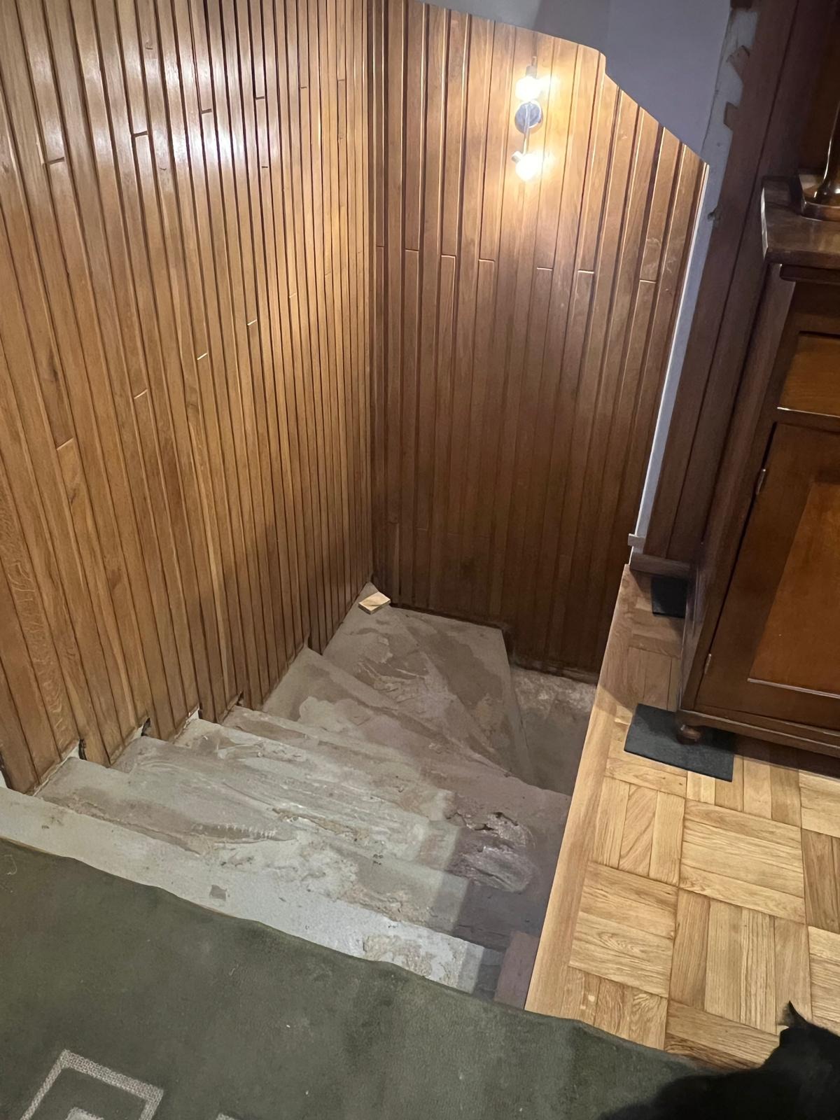 Schody w trakcie remontu, z drewnianą boazerią pionową na ścianach i widocznym oświetleniem. Fragment szafki i dywanu w dolnym rogu kadru.