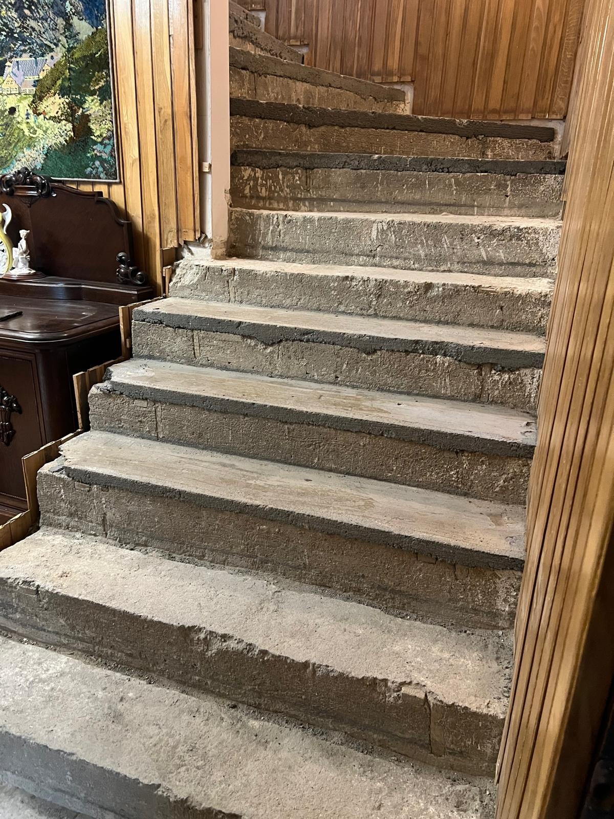Betonowe schody spiralne w trakcie remontu, z widocznymi śladami prac murarskich i fragmentami drewnianej boazerii w tle. Widoczne niedoskonałości i surowy stan wykończenia.
