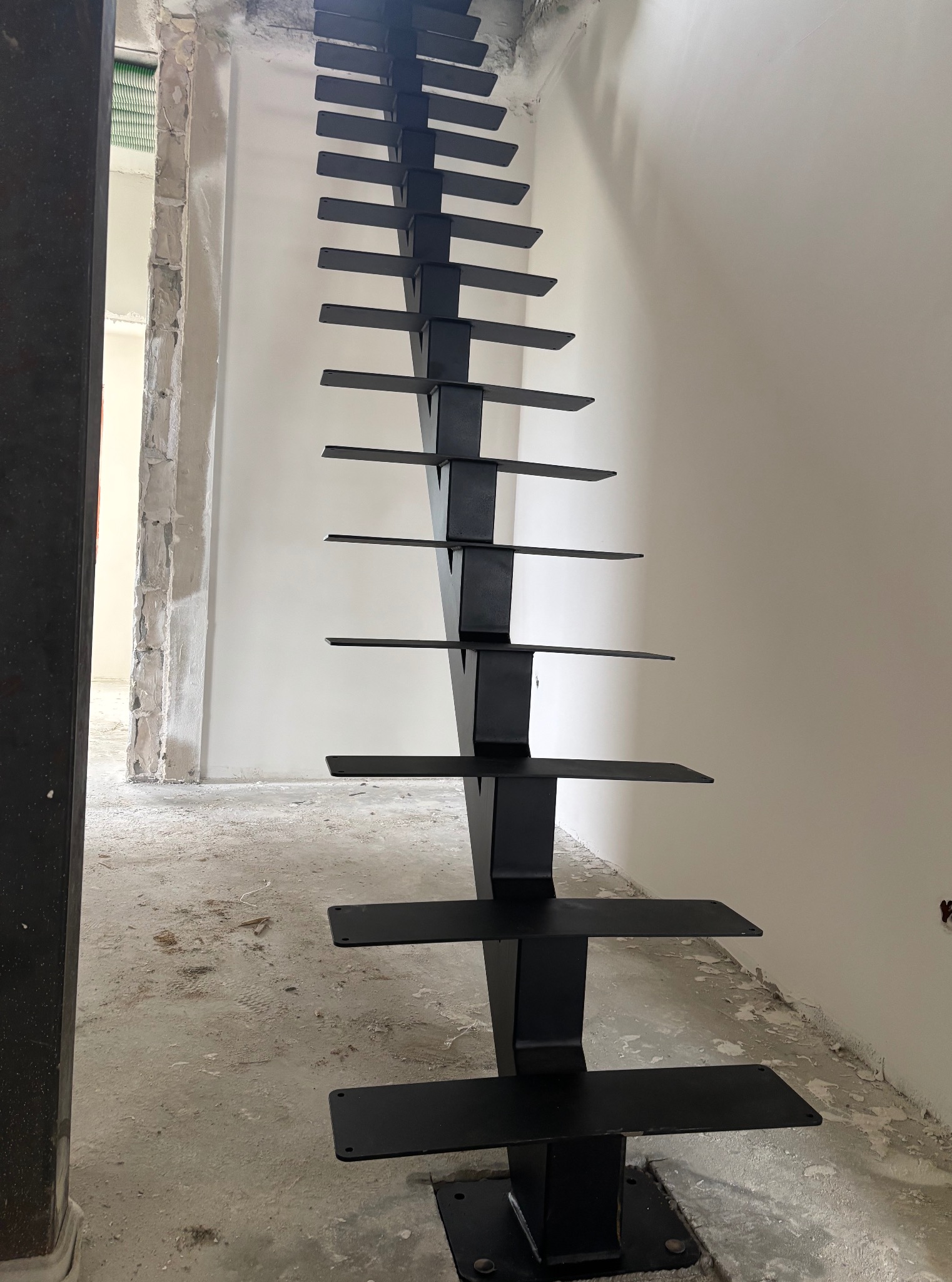 Surowe, czarne schody metalowe z prostymi stopniami, zamontowane w surowym wnętrzu w trakcie remontu. Widoczne niedokończone ściany i podłoga.