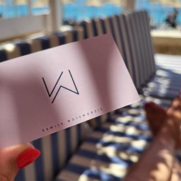 NOKO-CREATE PATRYK WOJEWODZIC - Różowa wizytówka z czarnym logo 'W' i imieniem Kamila Wojewodzic trzymana na tle plaży z leżakami.