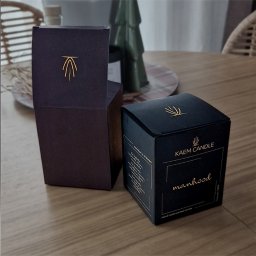 NOKO-CREATE PATRYK WOJEWODZIC - Dwa eleganckie, kwadratowe pudełka: otwarte, bordowe z złotym logo i zamknięte, czarne z napisem 'manhood' i logotypem 'KAEM CANDLE' w złotym kolorze.