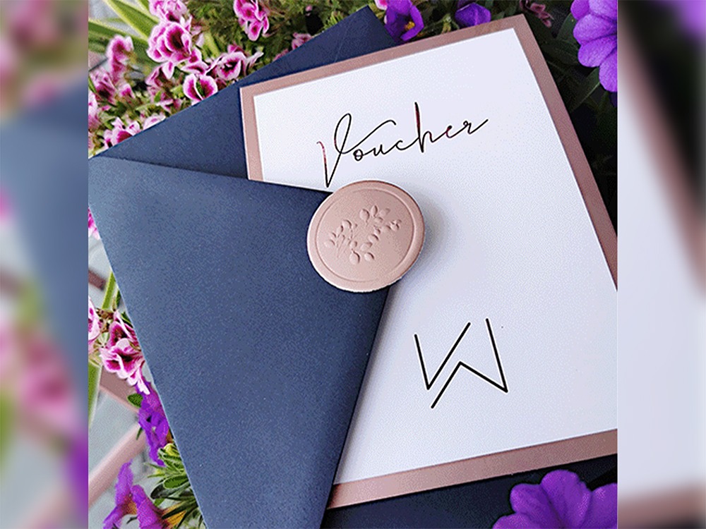 Elegancki voucher z pieczęcią lakową i granatową kopertą, otoczony kwiatami. Minimalistyczny design z napisem 'Voucher' i inicjałami.