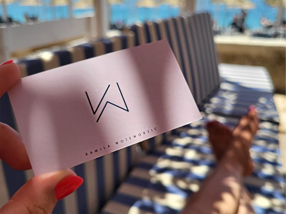 Różowa wizytówka z czarnym logo 'W' i imieniem Kamila Wojewodzic trzymana na tle plaży z leżakami.