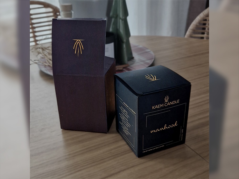 Dwa eleganckie, kwadratowe pudełka: otwarte, bordowe z złotym logo i zamknięte, czarne z napisem 'manhood' i logotypem 'KAEM CANDLE' w złotym kolorze.