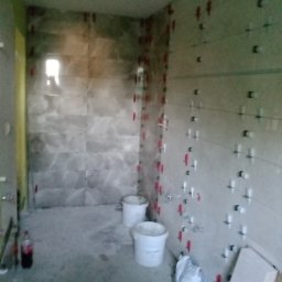 Igor Sobera - Łazienka w trakcie remontu: na ścianach ułożone płytki z użyciem klipsów poziomujących, na podłodze wiadra i materiały budowlane, butelka napoju.