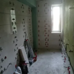 Łazienka w trakcie remontu: częściowo ułożone płytki na ścianach z użyciem klipsów poziomujących, widoczny worek z zaprawą i narzędzia glazurnicze.