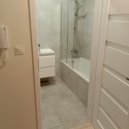 Igor Sobera - Nowoczesna łazienka z kabiną prysznicową nad wanną, umywalką z szafką i szarą płytką. Widok zza uchylonych, białych drzwi z poziomymi frezami.