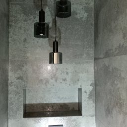 Igor Sobera - Nowoczesna łazienka z trzema szklanymi lampami wiszącymi na różnych wysokościach, wbudowaną wnęką i płytkami imitującymi beton, z minimalistycznym przyciskiem spłukującym.