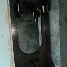 Igor Sobera - Owalne lustro w czarnej, fakturowanej ramie z dwoma cylindrycznymi lampami wiszącymi nad białą umywalką. Ściana wyłożona ciemnymi, kamiennymi płytkami.