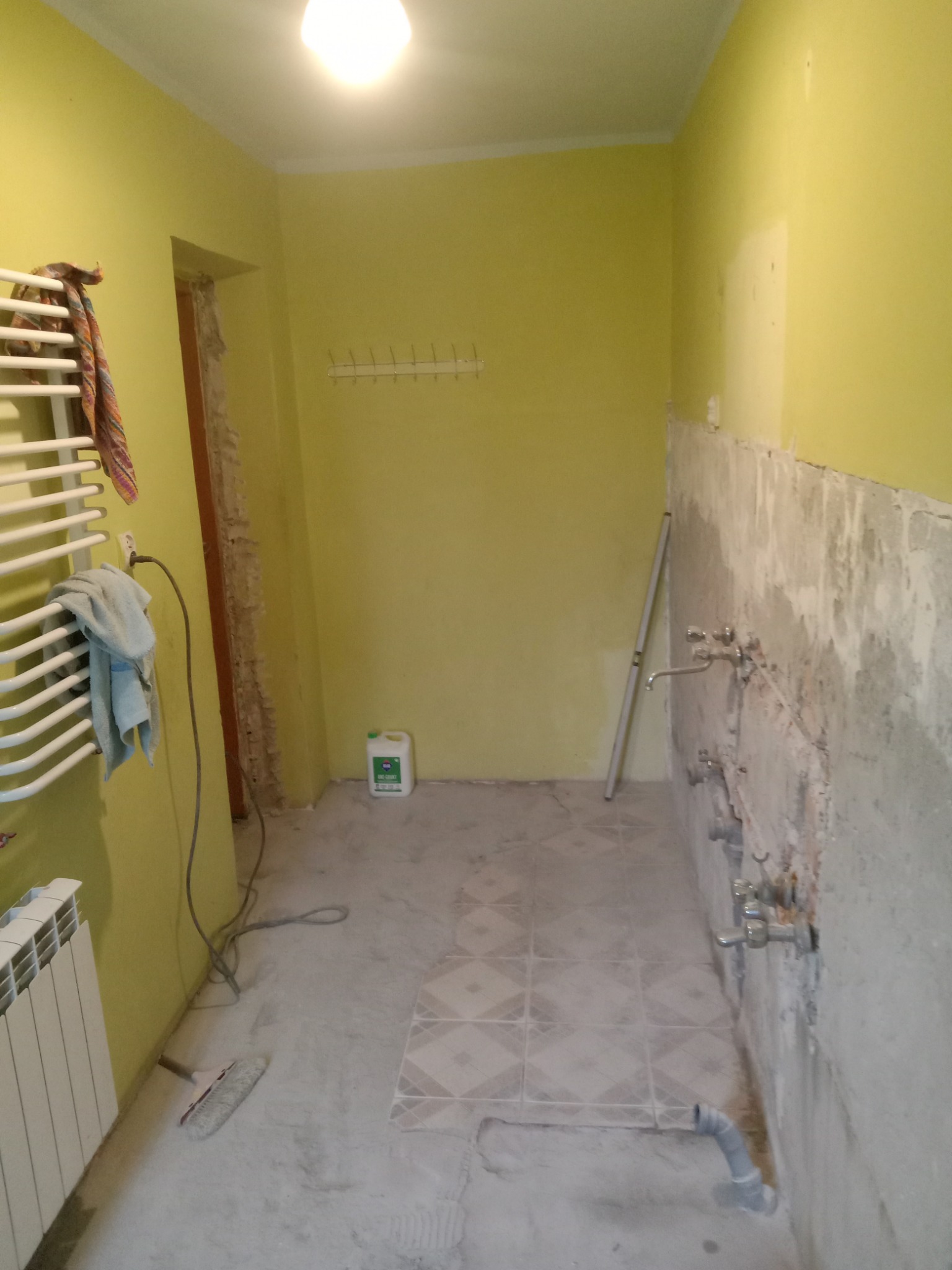 Żółta łazienka w trakcie remontu: odsłonięte rury, częściowo położone płytki podłogowe, grzejnik, poziomica oparta o ścianę, butelka chemii budowlanej.
