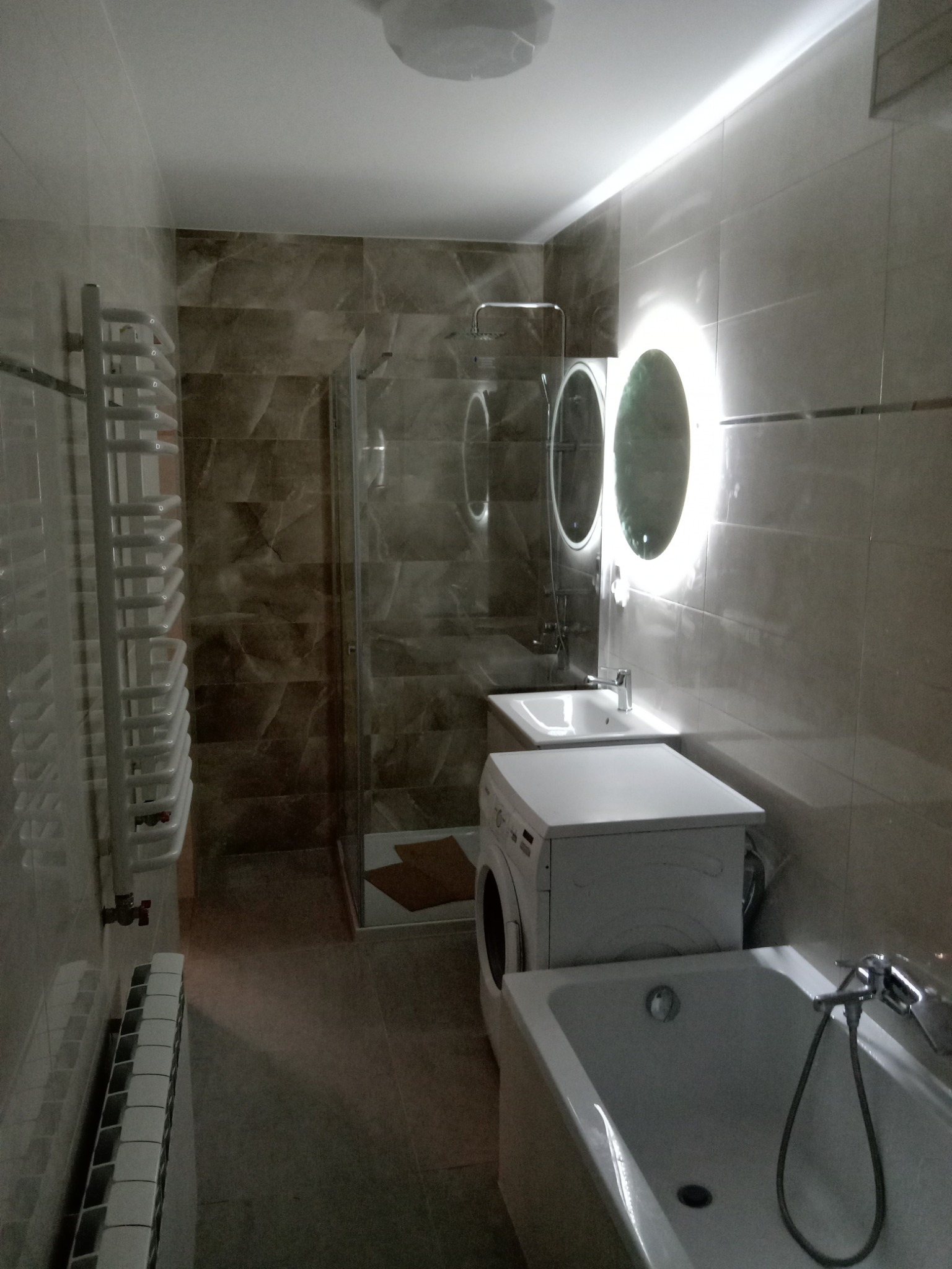 Wykończona łazienka z wanną, pralką zabudowaną pod umywalką, grzejnikiem drabinkowym i kabiną prysznicową z mozaiką w odcieniach beżu i brązu.