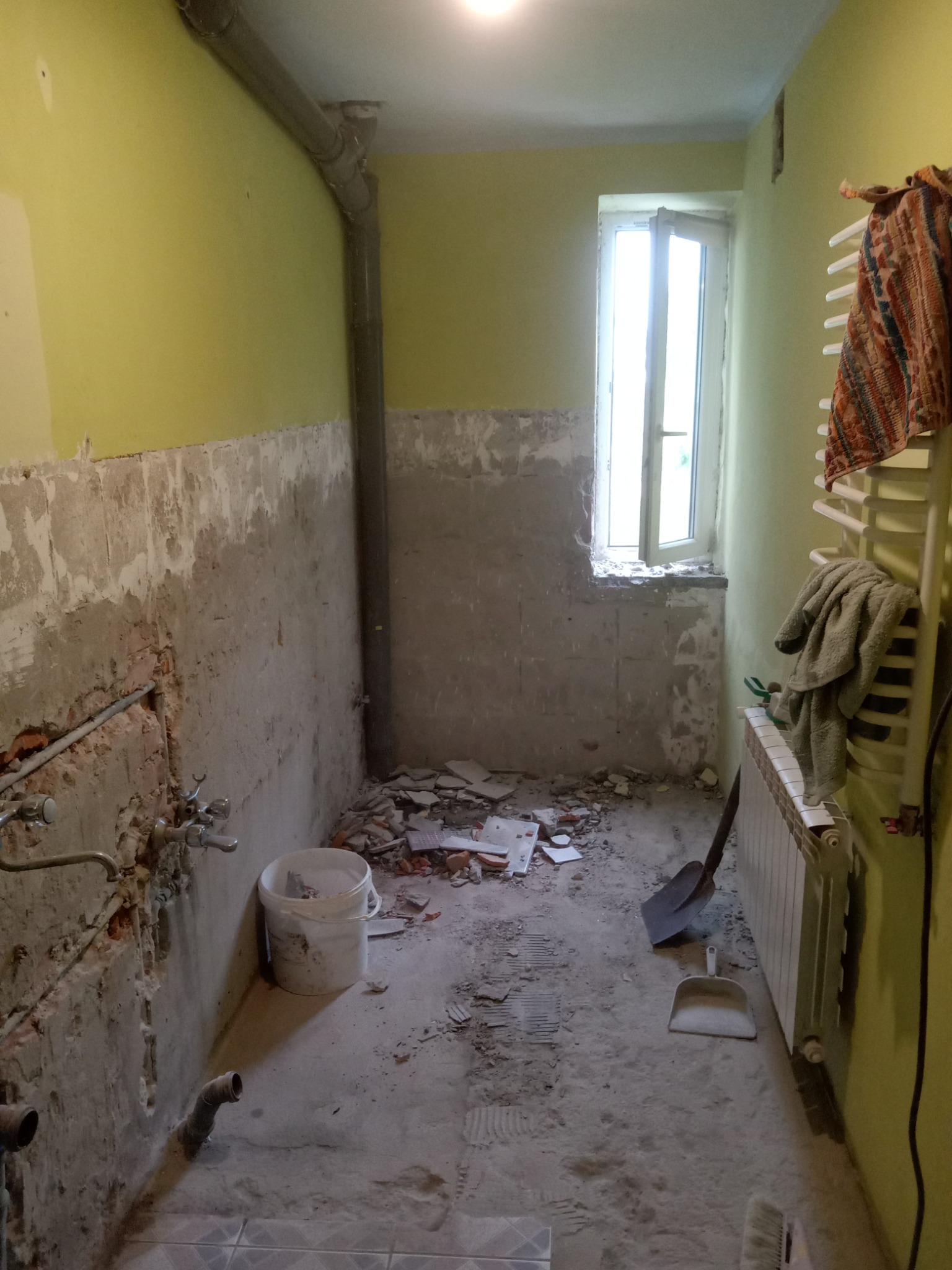 Łazienka w trakcie demontażu: odsłonięte rury, resztki płytek, żółte ściany, okno z naturalnym światłem. Widoczne narzędzia: łopata i szufelka.
