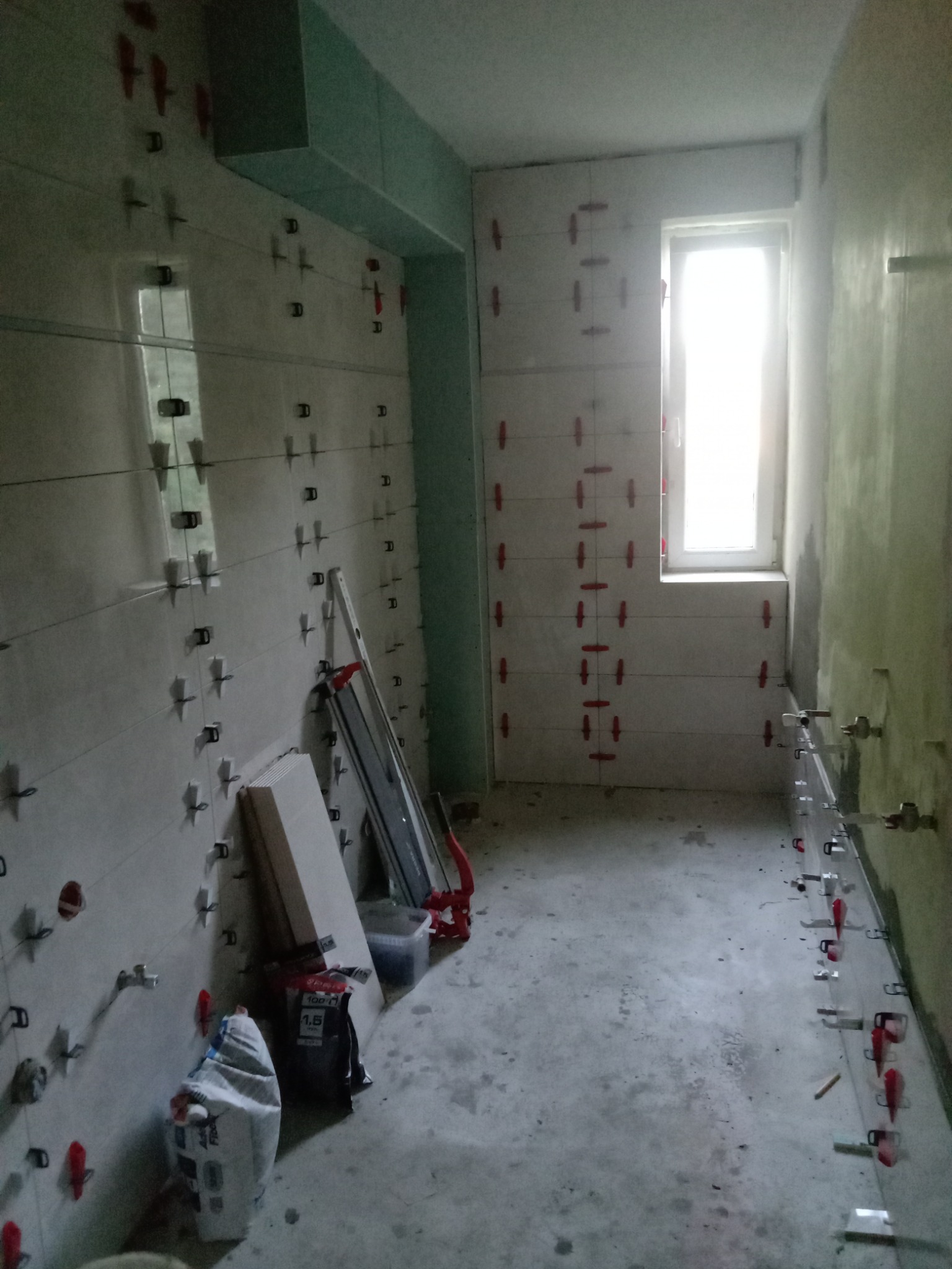 Łazienka w trakcie remontu: częściowo ułożone płytki na ścianach z użyciem klipsów poziomujących, widoczny worek z zaprawą i narzędzia glazurnicze.