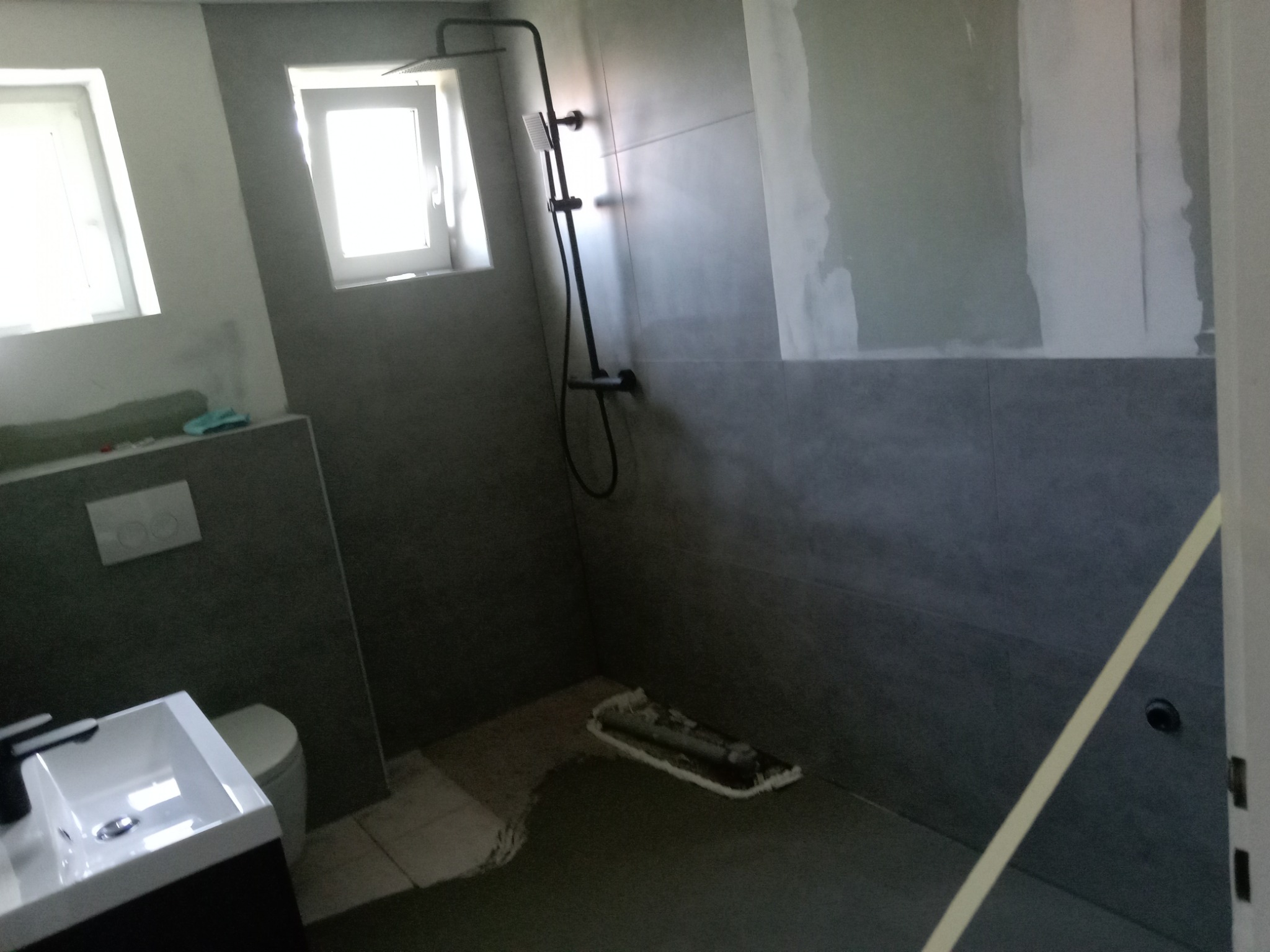 Szara łazienka w trakcie prac wykończeniowych: widoczna kabina prysznicowa, umywalka, okno i narzędzie na podłodze. Ściany częściowo wykończone.