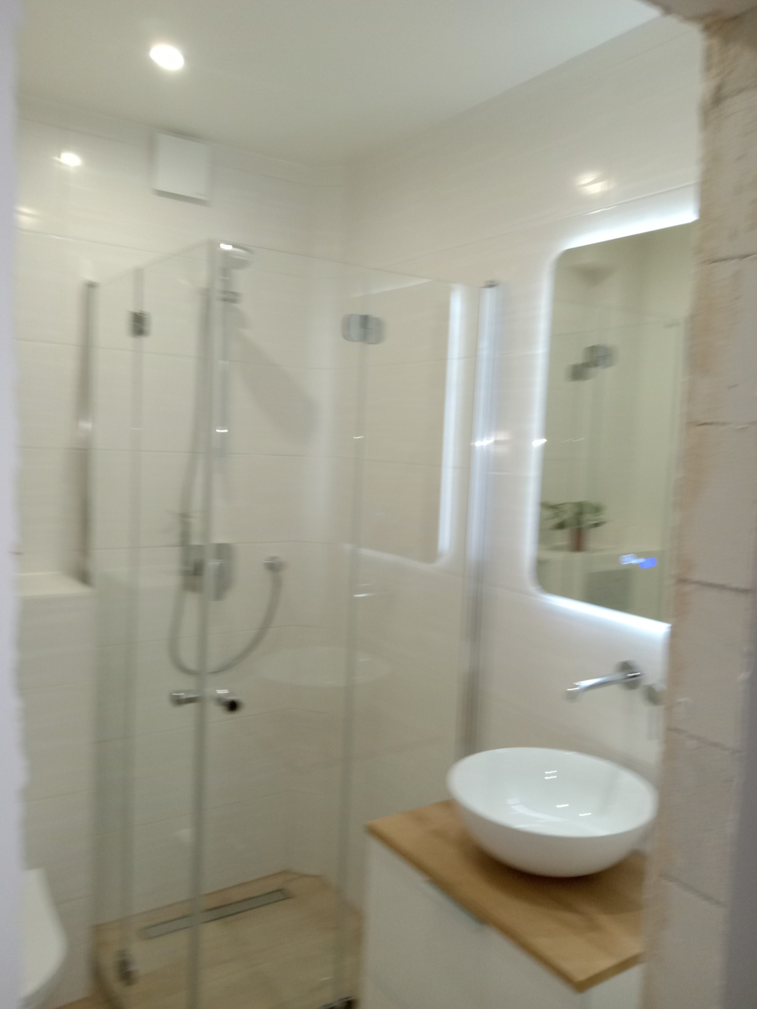 Nowoczesna łazienka z kabiną prysznicową typu walk-in, umywalką nablatową na drewnianym blacie i podświetlanym lustrem. Minimalistyczny design i jasna kolorystyka.