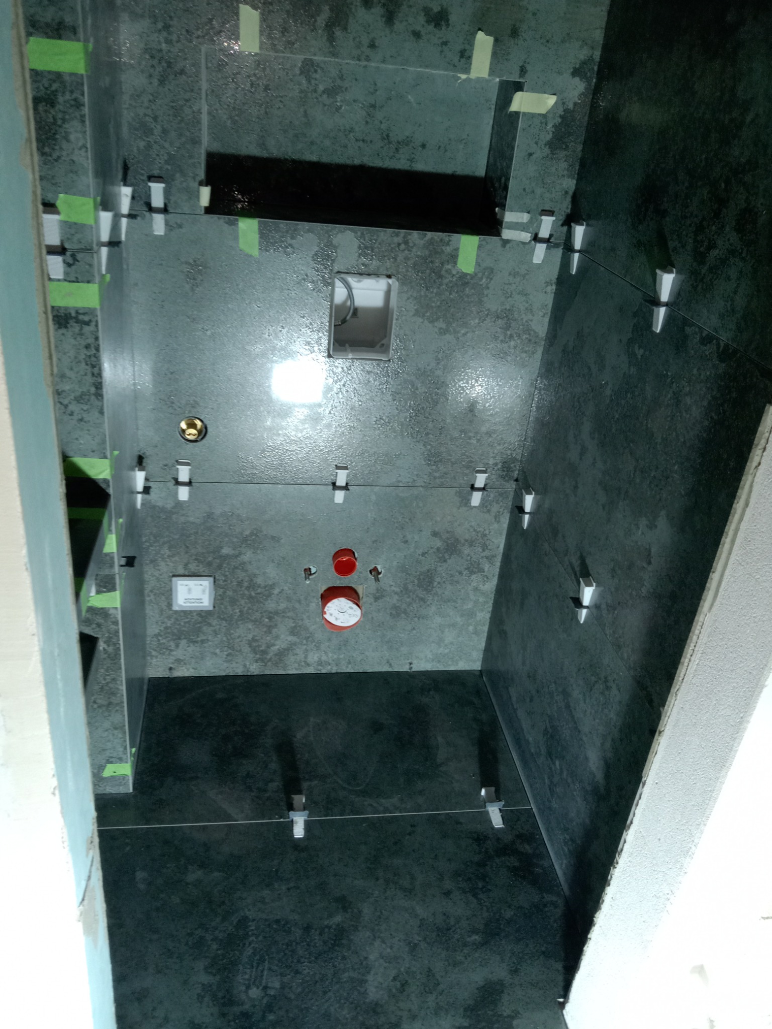 Ciemne płytki w trakcie układania w łazience, widoczne krzyżyki dystansowe i taśma maskująca, pionowy kadr z perspektywą od dołu.