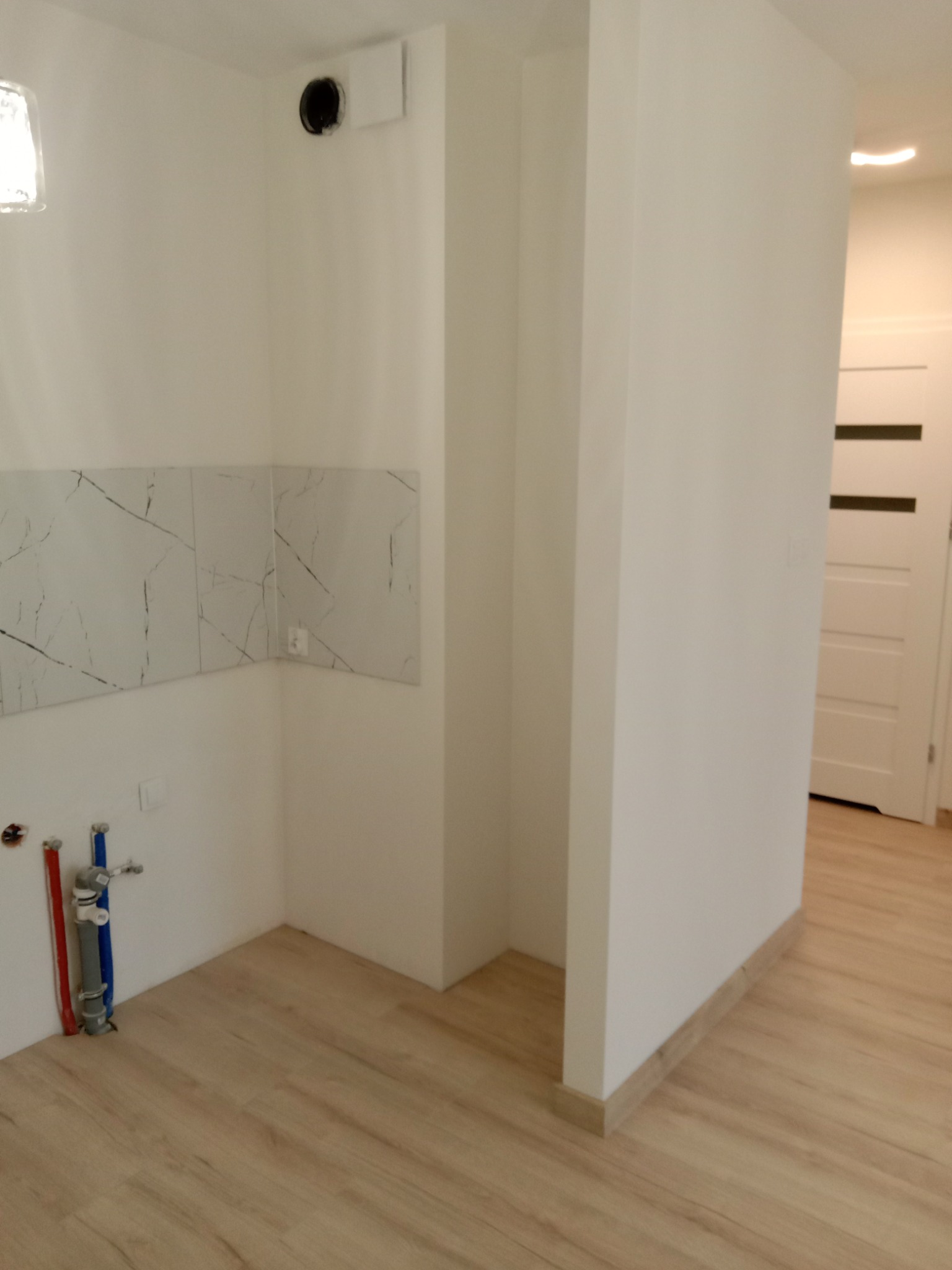 Wnętrze łazienki w trakcie wykańczania: białe ściany z marmurowymi płytkami, widoczne rury instalacyjne, jasna podłoga. Widok na otwarte przejście do innego pomieszczenia.