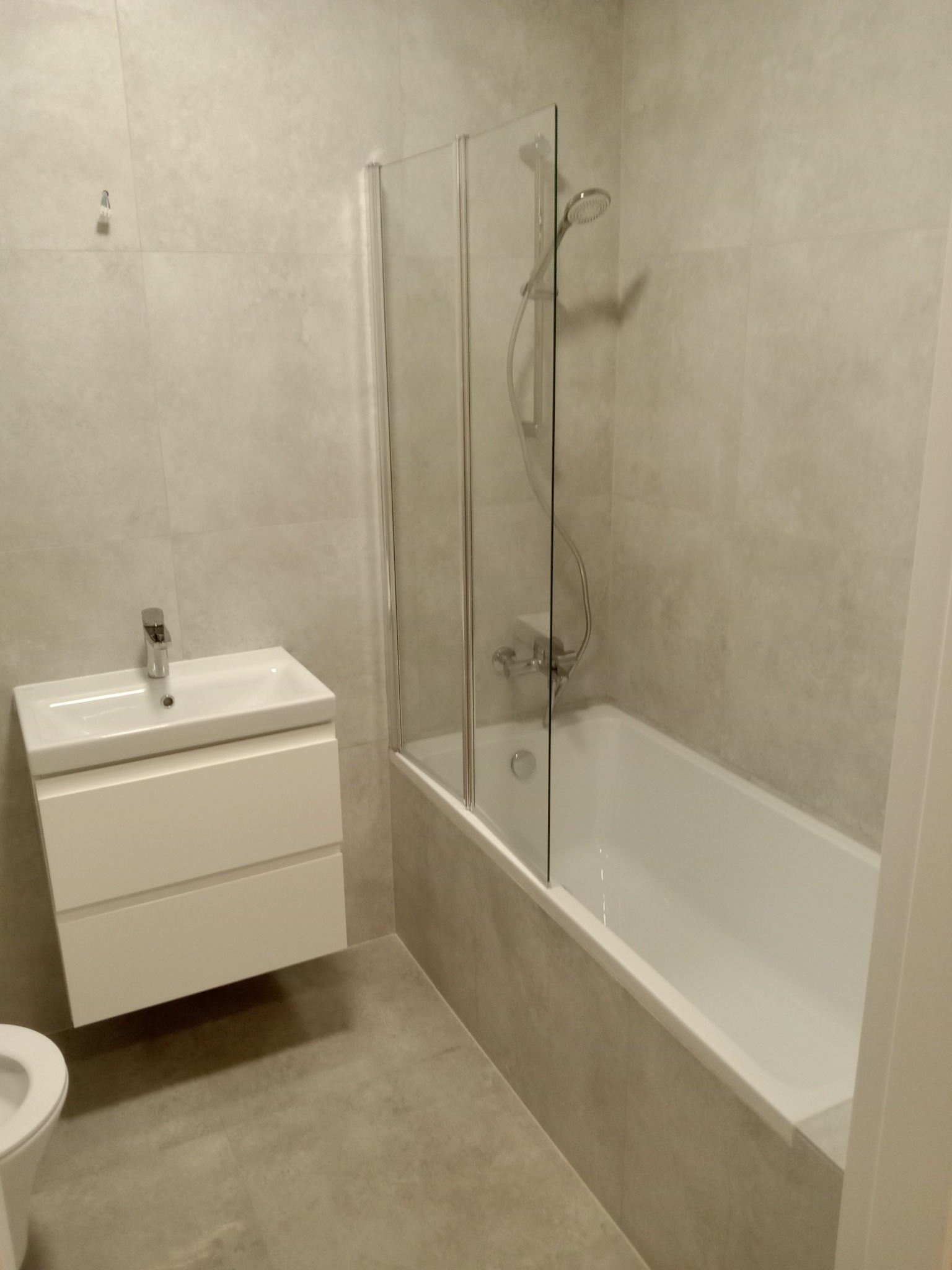 Jasna łazienka po remoncie: biała umywalka z szafką, wanna z parawanem, szare płytki na ścianach i podłodze. Minimalistyczny design, nowoczesne wykończenie.