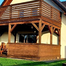 Timberland Pomorskie - Drewniana konstrukcja z balustradą przy domu jednorodzinnym, widok na zadbany trawnik. Elementy architektoniczne z ciemnego drewna. Balustrada z poziomych desek.