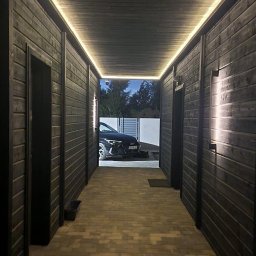 Timberland Pomorskie - Korytarz z ciemnego drewna z oświetleniem LED, prowadzący na zewnątrz, gdzie widoczny jest samochód. Podłoga wyłożona kostką brukową.  Kamera monitoringu na suficie.
