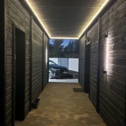 Korytarz z ciemnego drewna z oświetleniem LED, prowadzący na zewnątrz, gdzie widoczny jest samochód. Podłoga wyłożona kostką brukową.  Kamera monitoringu na suficie.