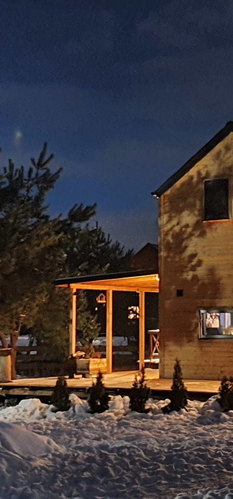 Zimowy wieczór: Drewniany dom z oświetlonym tarasem i pergolą. Śnieg leży na ziemi, a w oknach widać ciepłe światło. Widoczne drzewa iglaste.