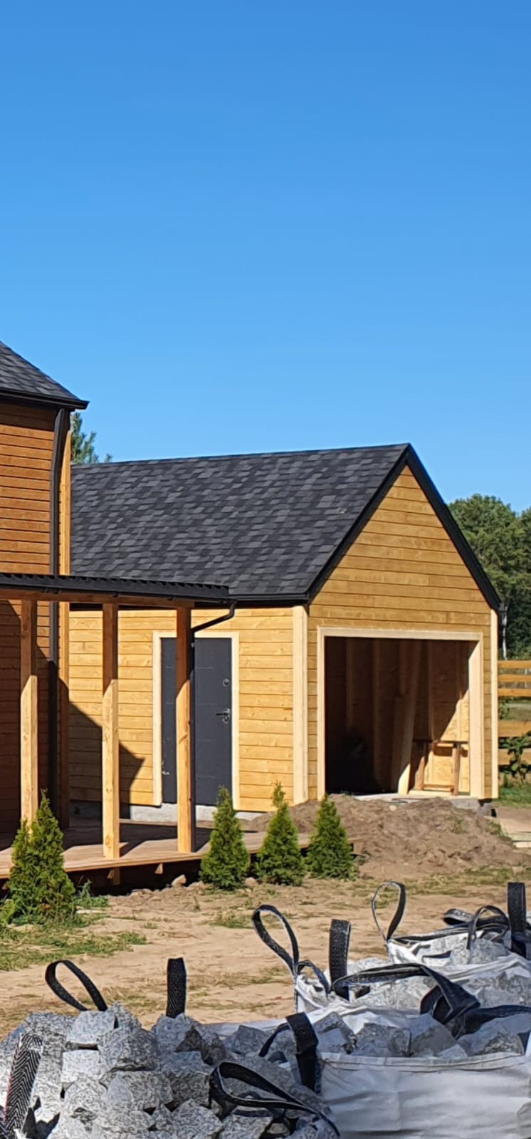 Drewniana konstrukcja w trakcie budowy z widoczną elewacją, dachem i elementami tarasu. W tle błękitne niebo i zieleń drzew. Na pierwszym planie worki z materiałem budowlanym.