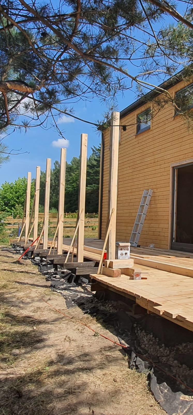 Budowa tarasu przy domu z desek. Widoczne słupy konstrukcyjne, czarna folia i częściowo ułożone deski. W tle drzewa i drabina oparta o ścianę budynku.