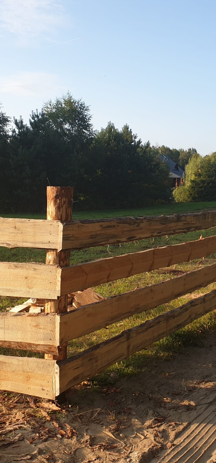 Surowe drewniane ogrodzenie w rustykalnym stylu, z widocznym pniem drzewa jako słupkiem. W tle zieleń drzew i budynek z ciemnym dachem. Ujęcie w słoneczny dzień.