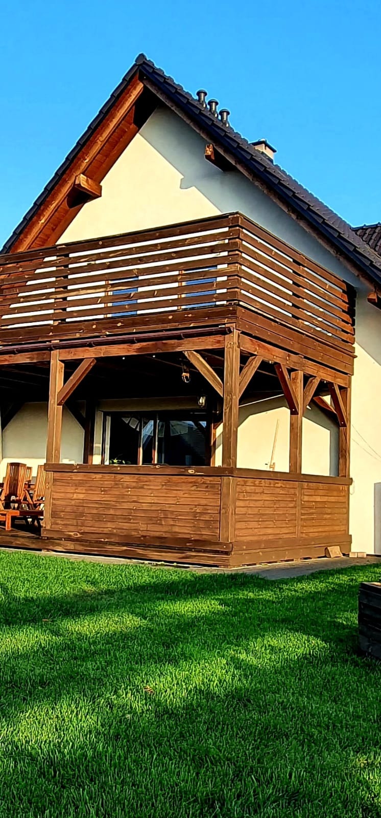 Drewniana konstrukcja z balustradą przy domu jednorodzinnym, widok na zadbany trawnik. Elementy architektoniczne z ciemnego drewna. Balustrada z poziomych desek.