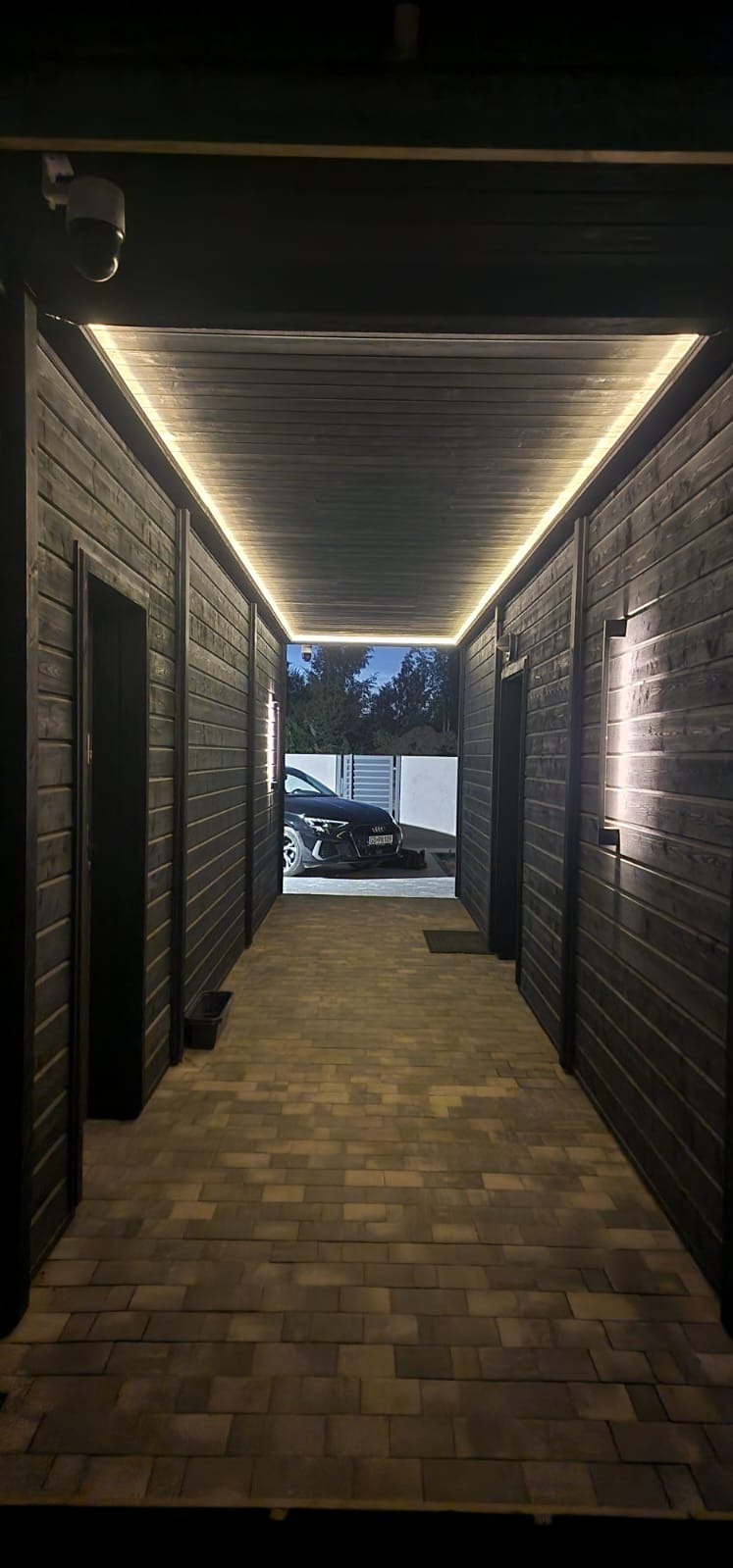 Korytarz z ciemnego drewna z oświetleniem LED, prowadzący na zewnątrz, gdzie widoczny jest samochód. Podłoga wyłożona kostką brukową.  Kamera monitoringu na suficie.