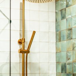 OiKa Studio Aleksandra Kośmider - Złota bateria prysznicowa z deszczownicą na tle białych i turkusowych płytek w łazience. Widoczny fragment szklanego parawanu.