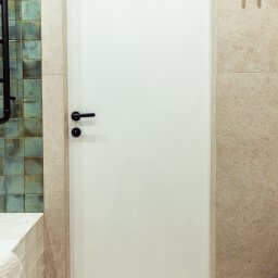 OiKa Studio Aleksandra Kośmider - Minimalistyczna, biała łazienkowa z czarną klamką, wkomponowana w beżowe płytki. Z lewej strony widoczne turkusowe akcenty i fragment grzejnika.